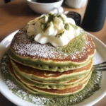 Matcha pancakes stack dusted with powdered sugar and topped with whipped cream and matcha powder, served alongside coffee
