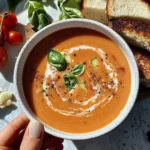 Overhead view of creamy tomato soup in a white speckled bowl garnished with fresh basil leaves and cream swirl, served with toasted bread and fresh tomatoes.
