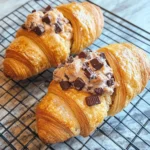 Close-up of two Le Crookie Cookie Croissants on a cooling rack, showing flaky pastry layers and cookie centers topped with chocolate pieces.