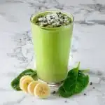 Green power smoothie in tall glass with coconut flakes, cacao nibs, banana slices and fresh spinach leaves on marble background