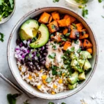 Top-down view of a Sweet Potato Rice Bowl with roasted sweet potatoes, diced avocado, black beans, red onion, cilantro, and creamy sauce on a bed of rice with corn.