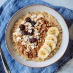Quinoa breakfast bowl topped with sliced banana, blackberries, coconut flakes, chopped nuts, and almond butter swirl on a white ceramic plate with blue napkin