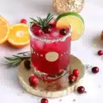 Glass of bright red cranberry mocktail with a coarse sugar rim, topped with fresh cranberries, a rosemary sprig, and a lime wheel on a gold coaster.