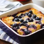 Square baking dish of Blueberry Buttermilk Pancake Casserole with abundant fresh blueberries on top, dusted with powdered sugar, on striped kitchen towel