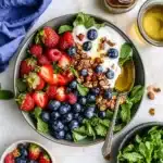 Fresh summer berry breakfast bowl featuring strawberries, blueberries, raspberries, yogurt, granola, and mint leaves arranged in a gray ceramic bowl with honey visible on the side.