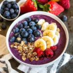 Purple acai smoothie bowl with organized toppings including blueberries, sliced banana, raspberries, and granola on a striped napkin.