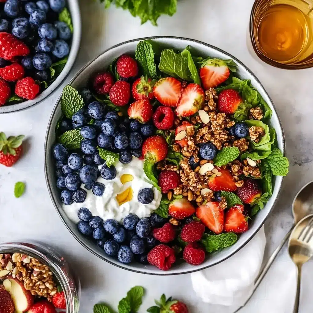Mixed berry breakfast bowl featuring blueberries, strawberries, raspberries, yogurt, granola, and mint leaves in a gray ceramic bowl with honey and additional ingredients surrounding it.