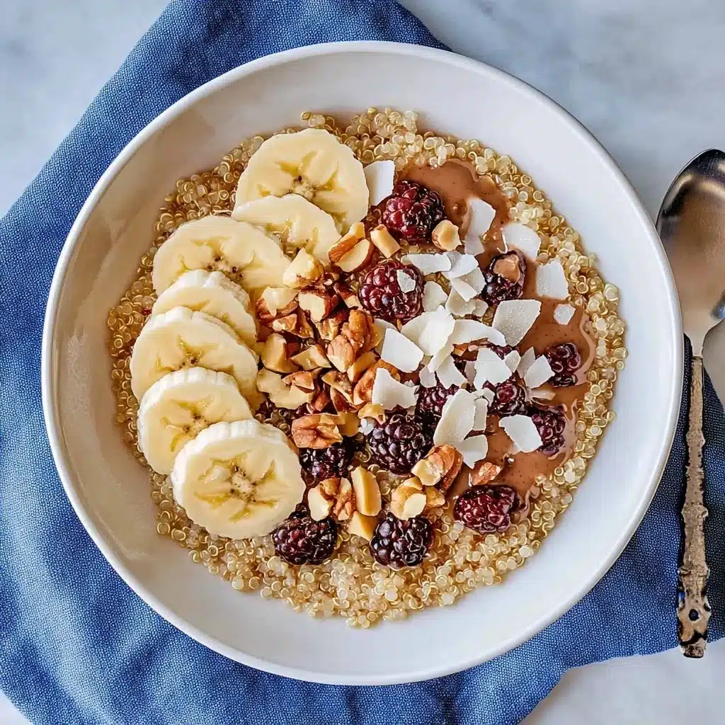 Overhead view of quinoa breakfast bowl with sliced banana, blackberries, chopped nuts, coconut flakes, and nut butter on white ceramic bowl with blue napkin