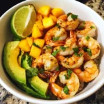 A white bowl with seasoned shrimp, mango chunks, avocado slices, and a lime wedge over a base of rice.
