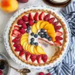 A beautiful Summer Fruit Tart on a decorative plate featuring strawberries, raspberries, blueberries and peach slices arranged in a colorful pattern atop a cream filling, with one slice removed.