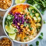 Thai Peanut Buddha Bowl featuring quinoa, purple cabbage, carrots, cucumber, avocado, cilantro, peanuts and peanut sauce with lime wedges