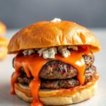 A close-up, high-angle view of a juicy, freshly made Buffalo blue cheese smash burger, set against a clean, light background. The burger bun is toasted to a golden brown, with a generous drizzle of buttery hot sauce cascading down the sides, creating a mouthwatering effect. The patty is seared to perfection, with crisp edges and a soft, tender center, topped with a melted blue cheese crumble. The overall composition conveys a sense of indulgence and the irresistible appeal of this flavorful, comfort-food creation.